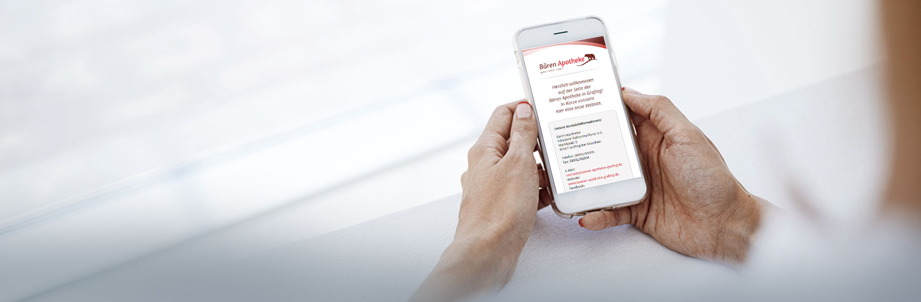 Zwei männliche Hände halten ein Handy mit Website der Bären Apotheke Grafing